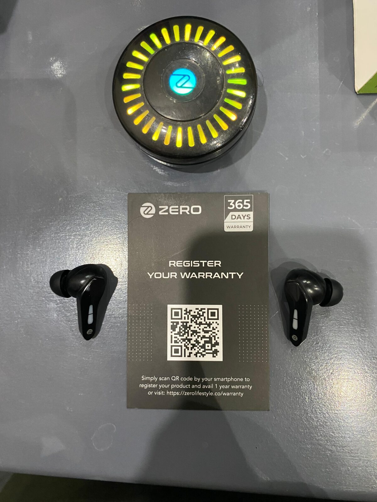 Zero Lifestyle Arcade 900 Z Buds 1-year warranty registration card with QR code, shown next to the glowing circular RGB charging case and earbuds.