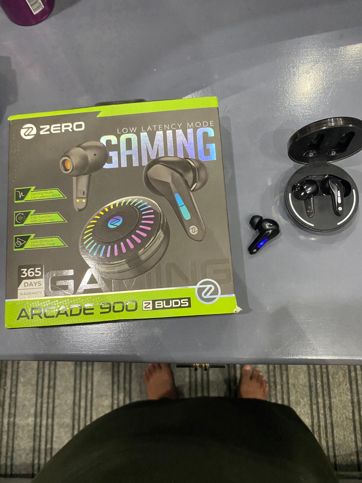 Zero Lifestyle Arcade 900 Z Buds retail box next to the open circular charging case and a black gaming earbud on a desk. earbud.
